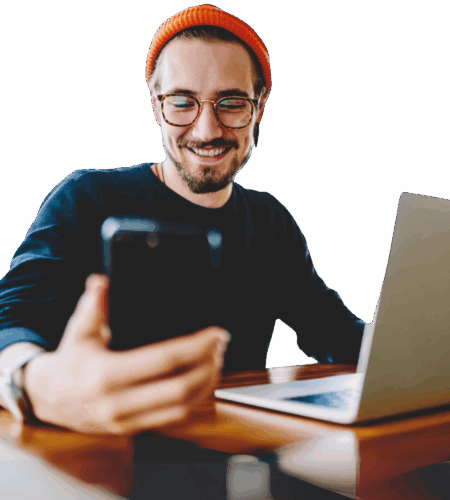 Ein Mann sitzt vor einem Laptop und sieht parallel auf sein Smartphone, das er in seiner Hand hält.