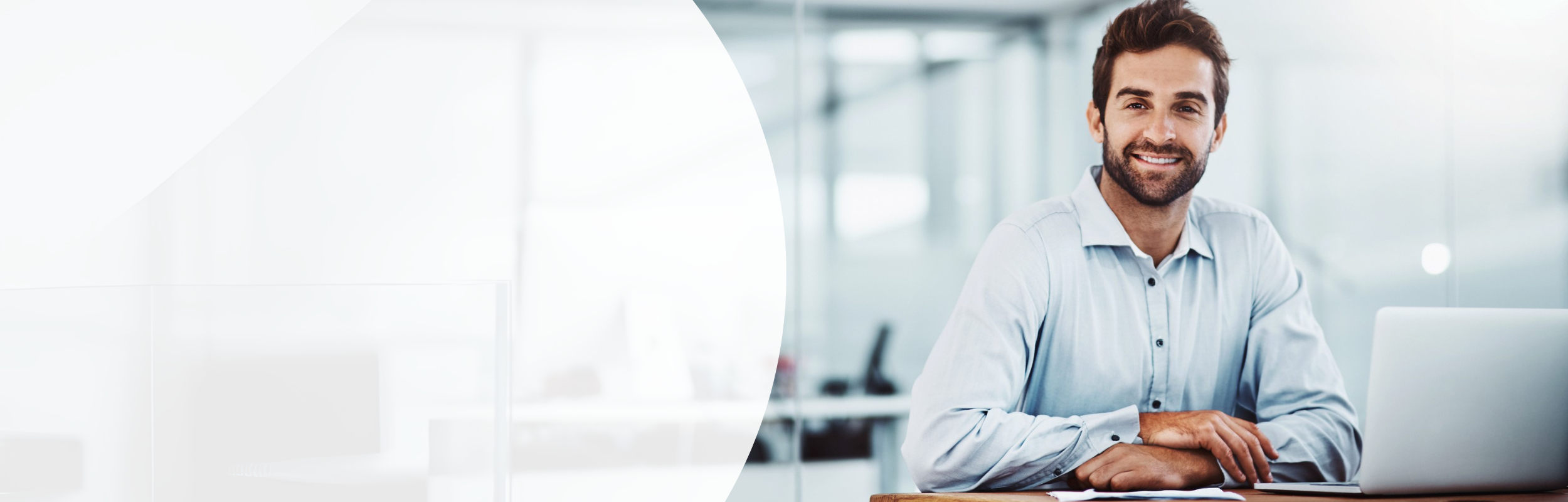 Mann in einem hellblauen Hemd sitzt mit verschränkten Händen vor seinem Laptop an einem Schreibtisch.