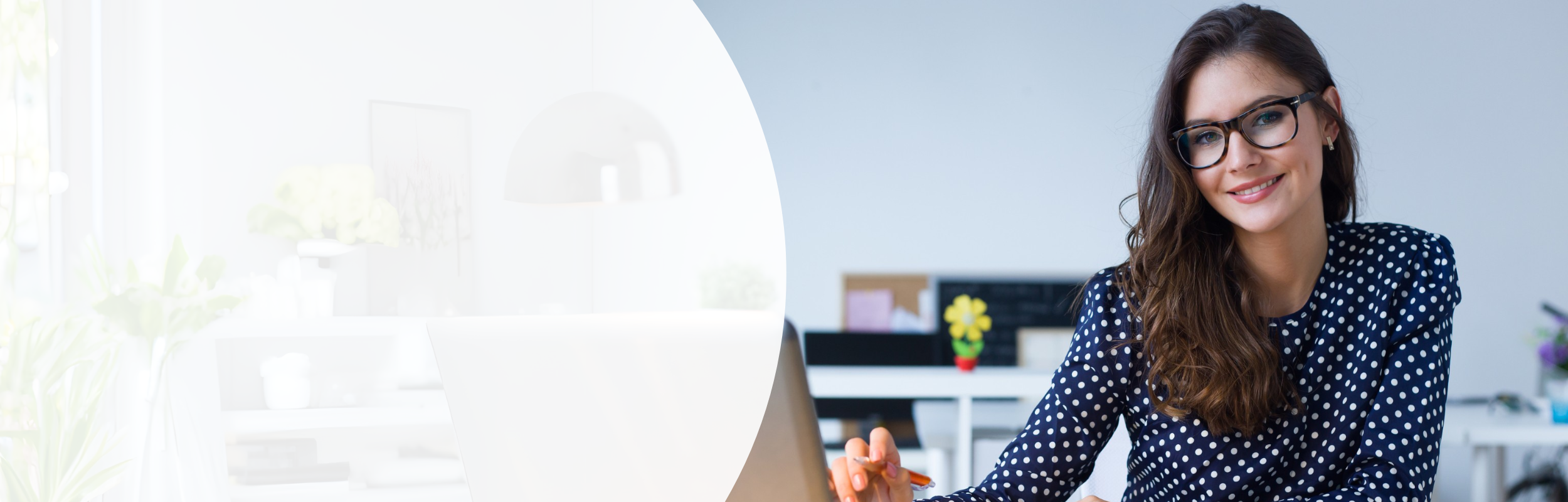 Frau mit langen braunen Haaren, einer gepunkteten Bluse und Brille sitzt vor Laptop