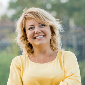 Portrait einer lächelnden Blonden Frau in einer gelben Bluse.