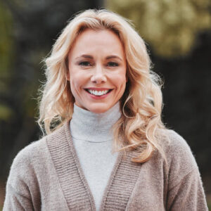 Portrait einer lächelnden Frau mit langen blonden Haaren, die einen grauen Pullover trägt, und eine braune Weste trägt.