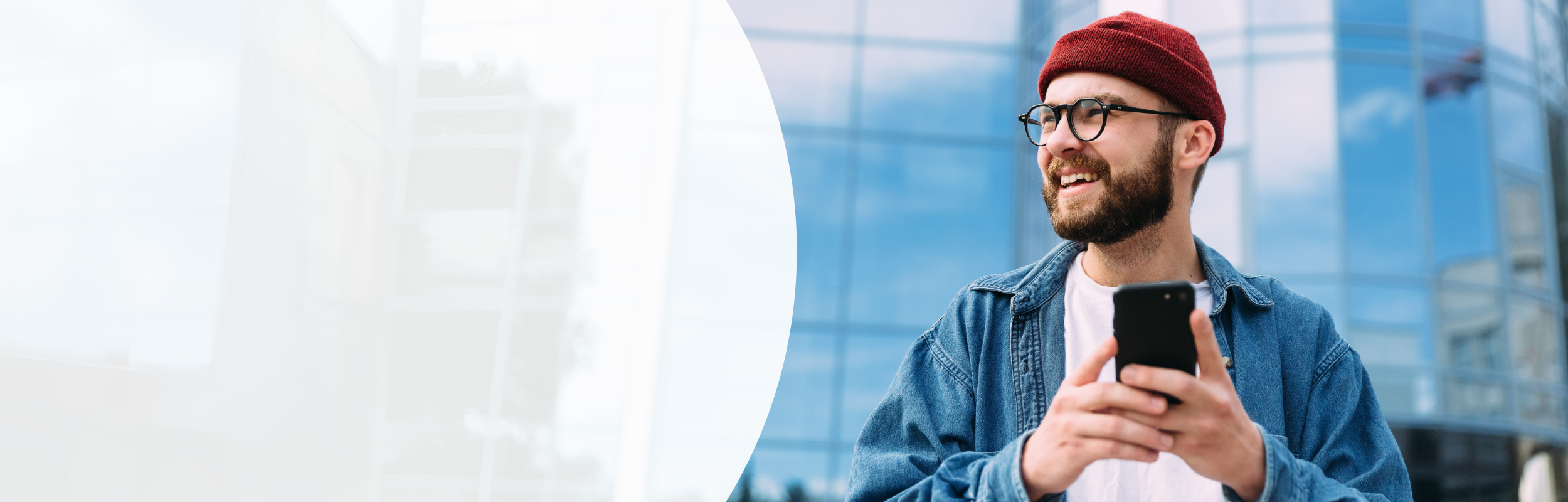 Mann mit Bart, roter Mütze und Brille mit Handy in der Hand vor einem Gebäude mit Glasfassade