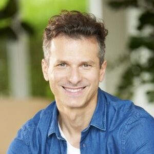 Ein Mann in blauem Hemd sitzt auf einer Couch und lächelt in die Kamera.