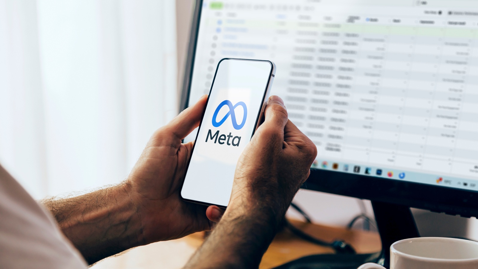 Social Media Werbung - Mann hält ein Smartphone, auf dem das Meta-Logo zu sehen ist. Im Hintergrund ein Computerbildschirm mit der Benutzeroberfläche des Meta Business Managers.