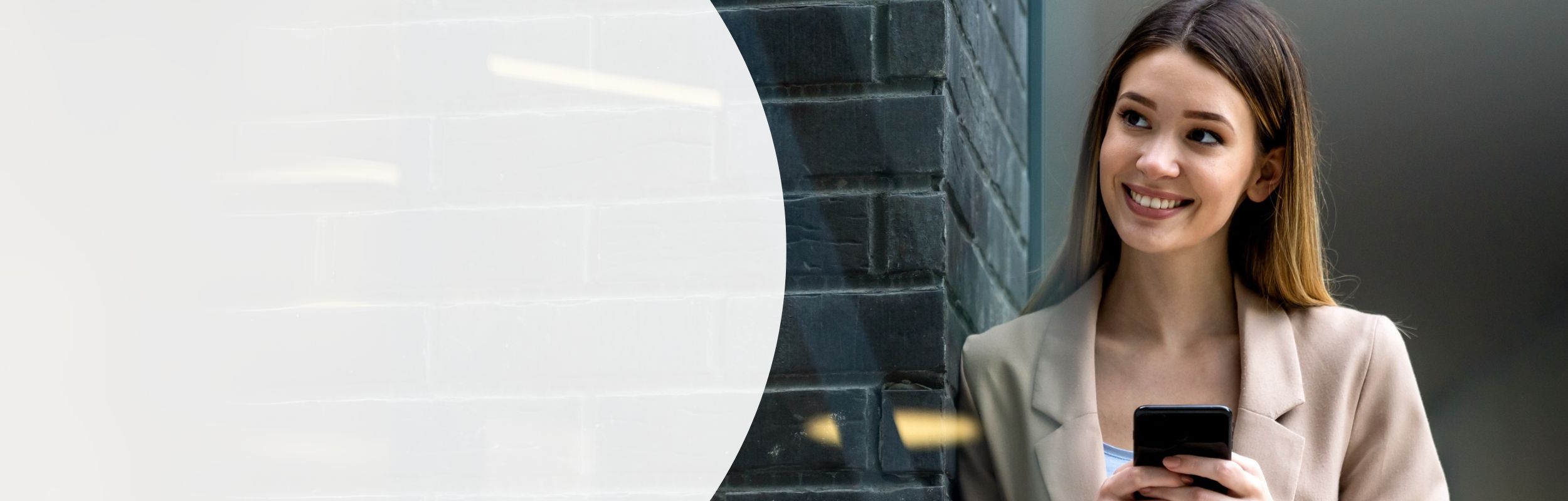 Lächelnde Frau mit langen braunen Haaren und einem beigen Blazer hält ein Smartphone in der Hand.