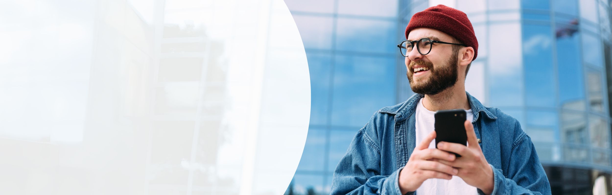 Mann mit Bart, roter Mütze und Brille mit Handy in der Hand vor einem Gebäude mit Glasfassade.