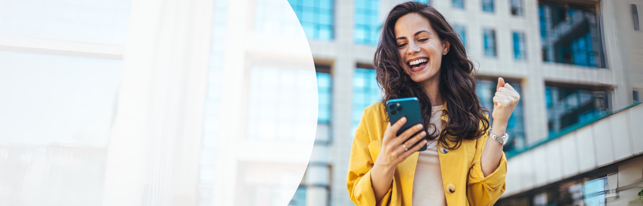 Frau mit langen brauen Haaren und gelber Jacke schaut voller Freude auf ihr Smartphone.
