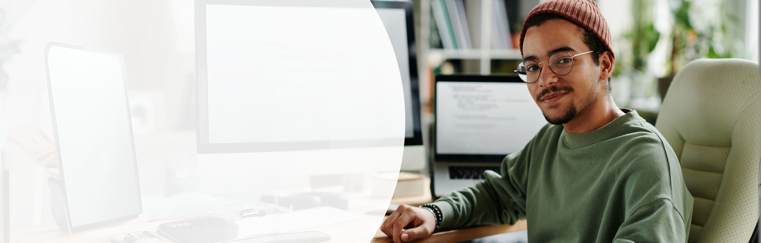 Ein Mann in einem dunkelgrünen Pullover und Mütze sitzt an einem Bildschirmarbeitsplatz.