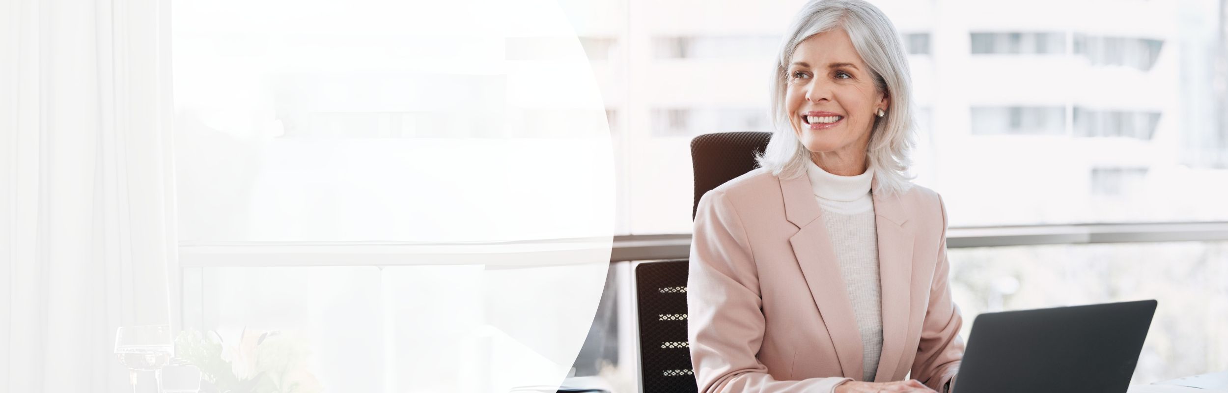 Lächelnde Frau mittleren Alters sitzt an einem Schreibtisch und hat einen schwarzen Laptop vor sich.