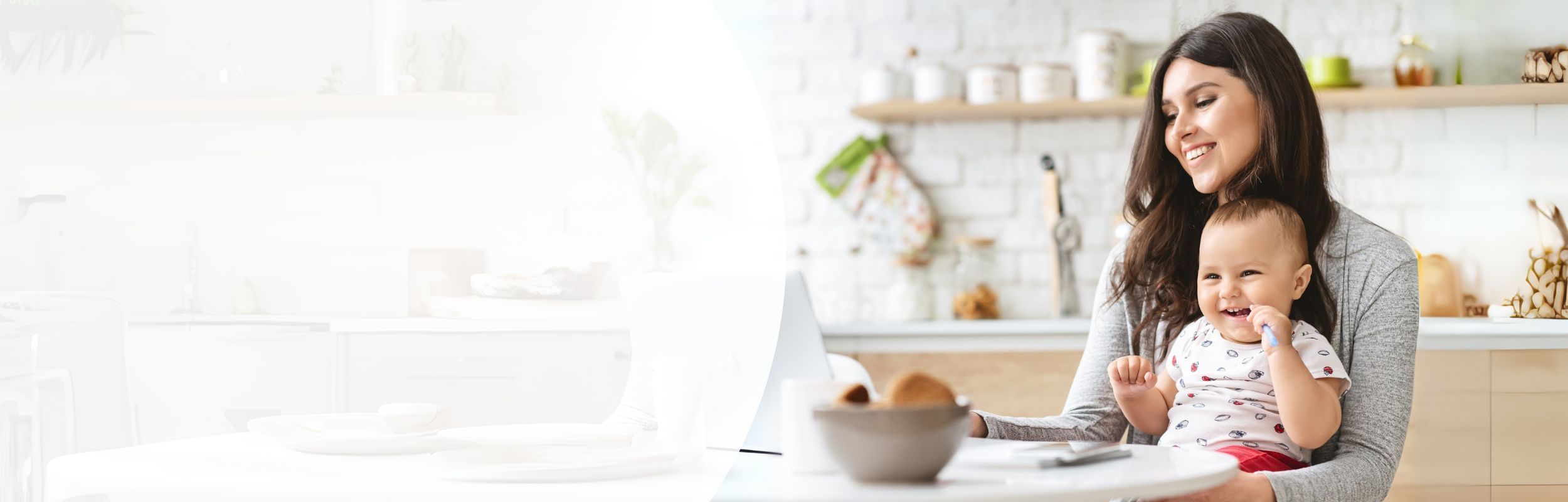 Frau sitzt in der Küche mit Baby auf dem Schoss vor einem Laptop.