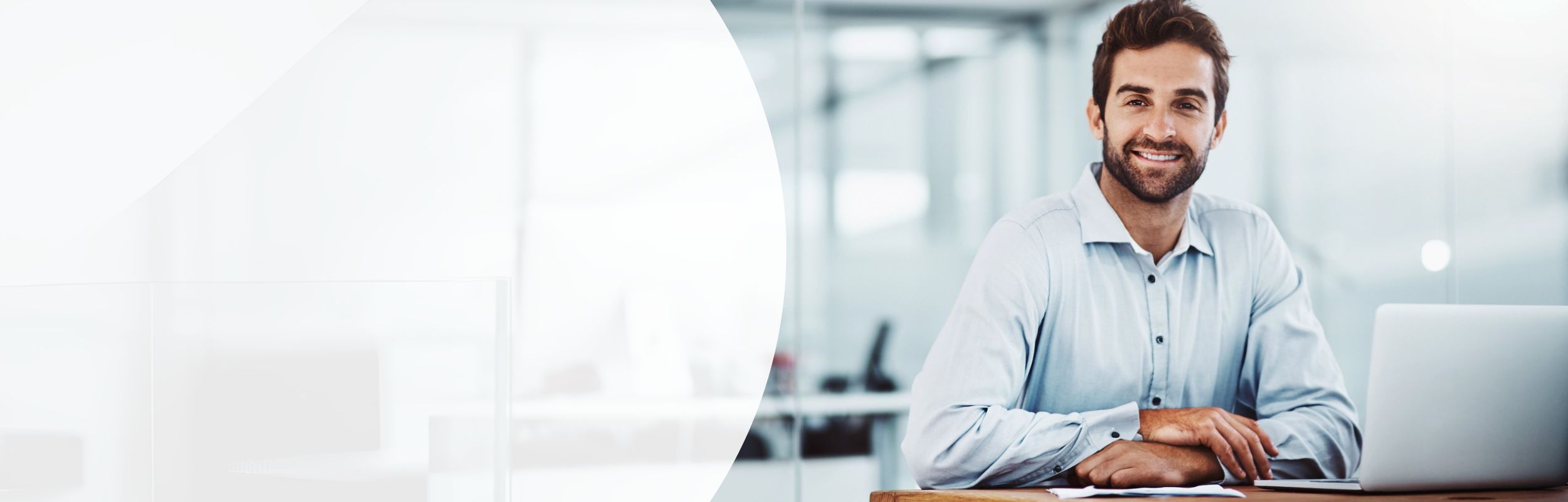 Ein Mann in hellblauem Hemd sitzt mit verschränkten Händen an einem Schreibtisch mit Laptop.