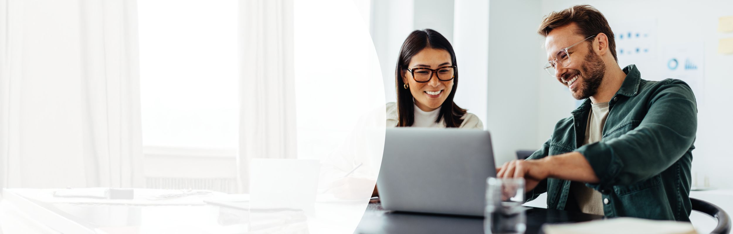 Ein Mann und eine Frau sitzen in geschäftlichem Kontext vor einem Laptop. Der Mann zeigt auf den Bildschirm.