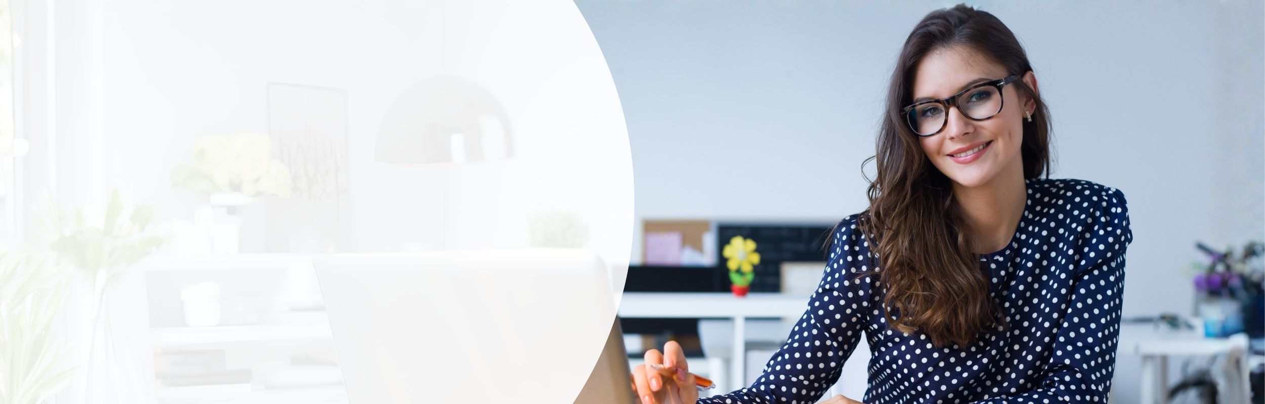 Frau mit Brille und blau-weiß gepunkteter Bluse sitzt an einem Schreibtisch mit Laptop.