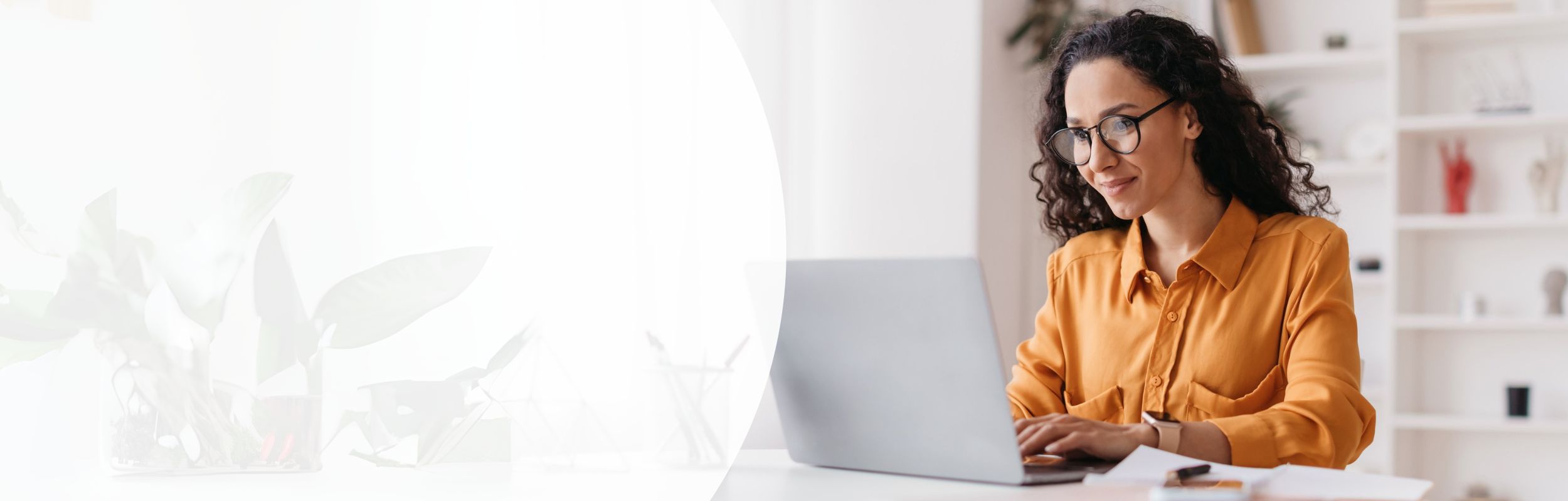 Frau mit langen gewellten Haaren, Brille und orangener Bluse sitzt vor einem Laptop.