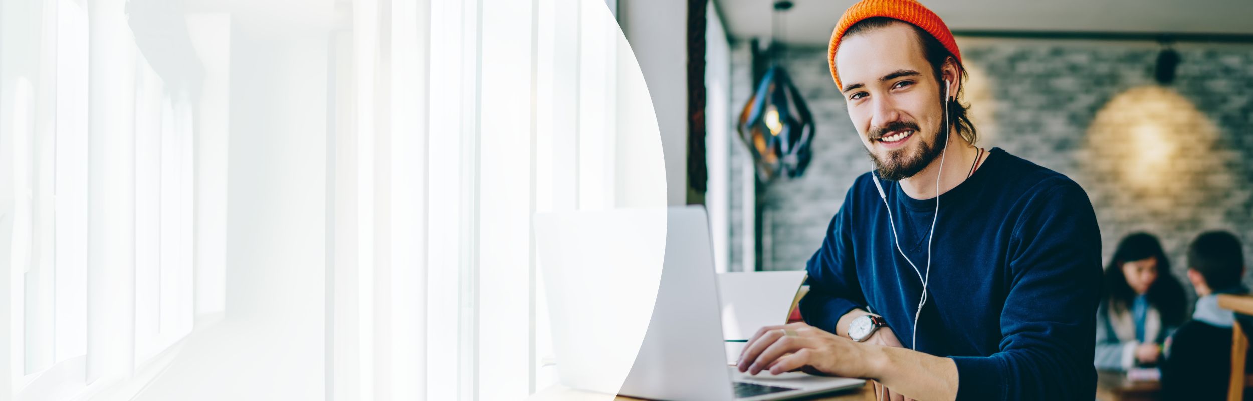 Mann mit Bart, Earphones und orangener Mütze sitzt an einem Laptop.