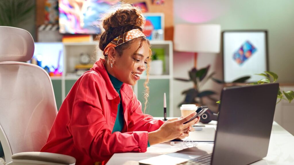 Junge Frau an einem Büroarbeitsplatz blickt auf ihr Smartphone.