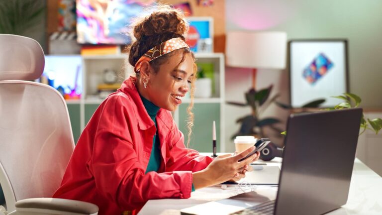Junge Frau an einem Büroarbeitsplatz blickt auf ihr Smartphone.