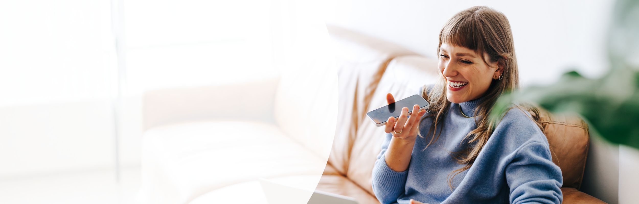 Frau mit langen braunen Haaren sitzt lachend auf einer Couch und spricht in ein Handy