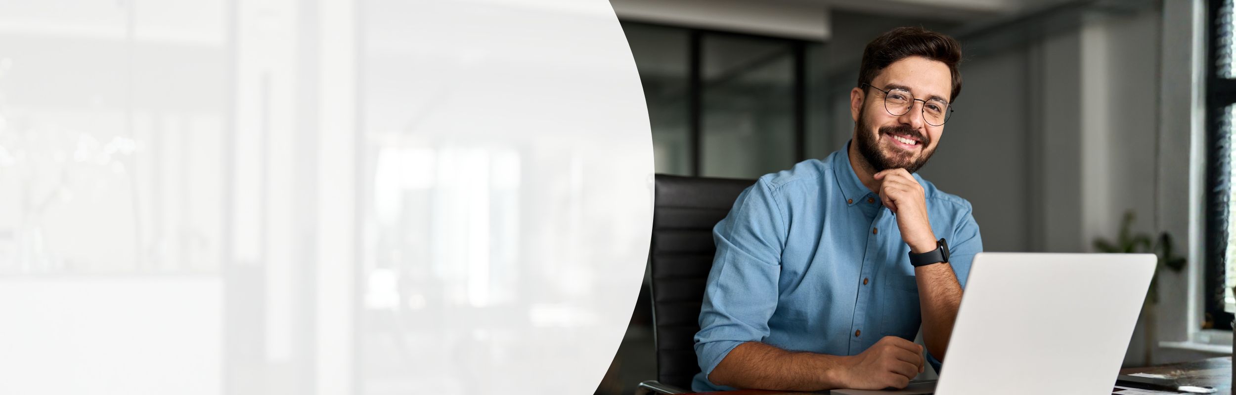Mann mit blauem Hemd, Bart und Brille sitzt lächelnd an seinem Laptop