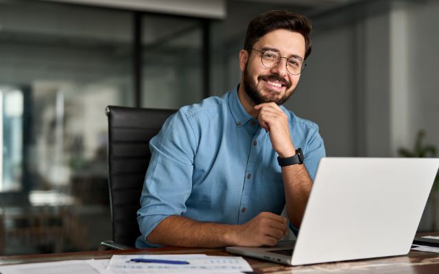 Mann mit blauem Hemd, Bart und Brille sitzt lächelnd an seinem Laptop