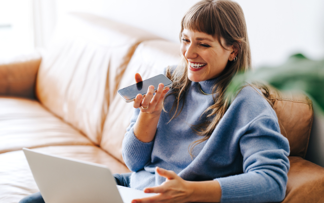 Frau mit langen braunen Haaren sitzt lachend auf einer Couch und spricht in ein Handy