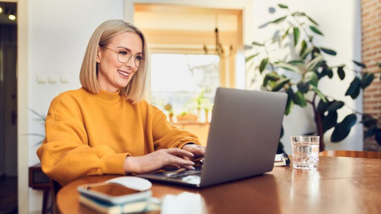 Social Media Selbstständig - Porträt einer Frau mittleren Alters, die im Esszimmer sitzt und mit einem Laptop zu Hause arbeitet.