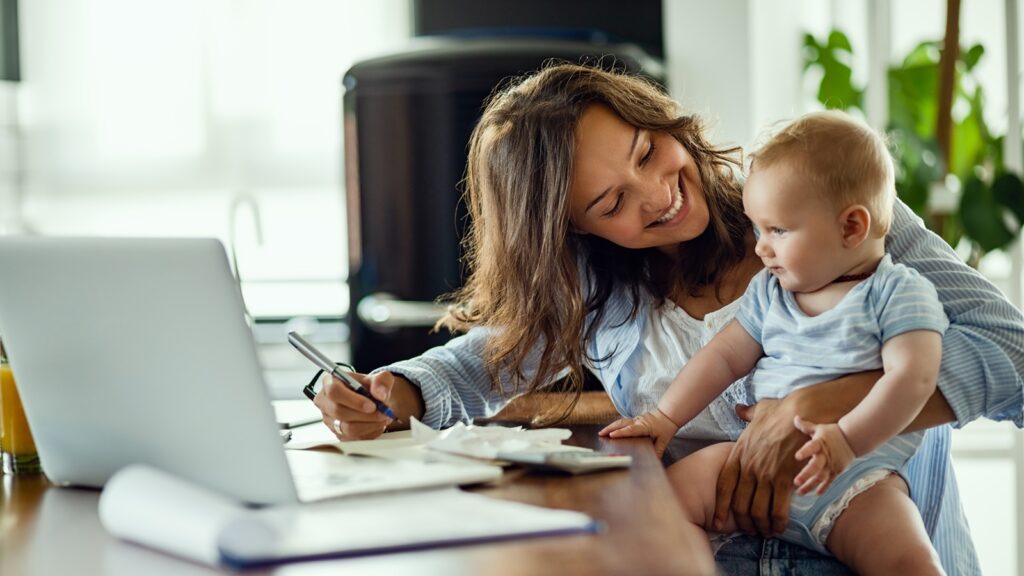 Rückkehr Elternzeit - Glückliche Mutter hält während des Arbeitens ihr Baby auf dem Arm.